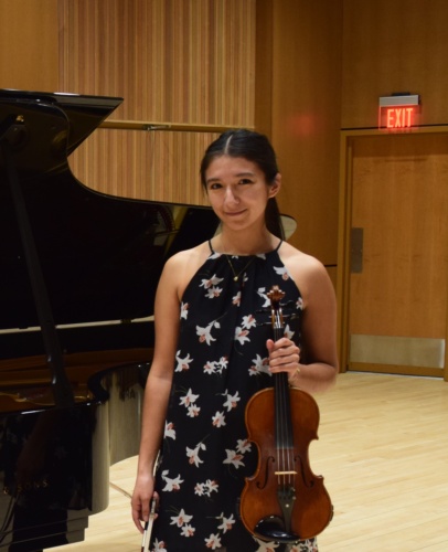 Raquel holding a violin, a piano behind her